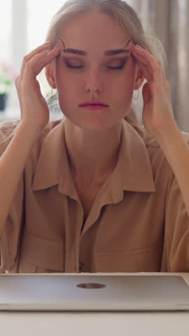 Tired young woman having headache sits by closed laptop at white table massaging weary forehead with hands against window in room slow motion