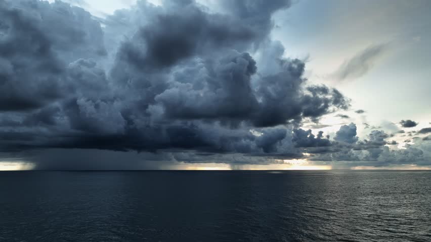 A drone captures the dark, swirling clouds above the ocean, as an intense rainstorm begins to form, with rain visible in the distance, signaling a powerful weather event.

