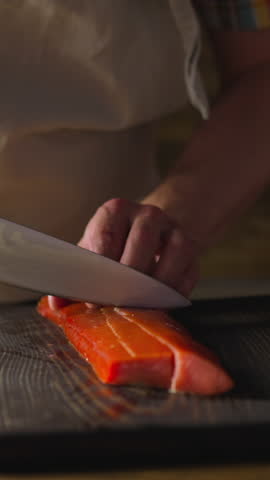 Employee in apron cuts delicious cool smoked chum salmon fillet into pieces with knife on wooden board at countertop closeup slow motion. Cooking seafood