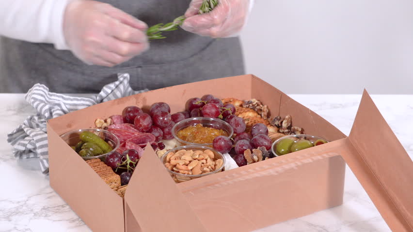Hands are meticulously adding fresh red grapes to a bowl, complementing a beautifully arranged charcuterie box brimming with a variety of cheeses, olives, and cured meats, set against a sleek marble