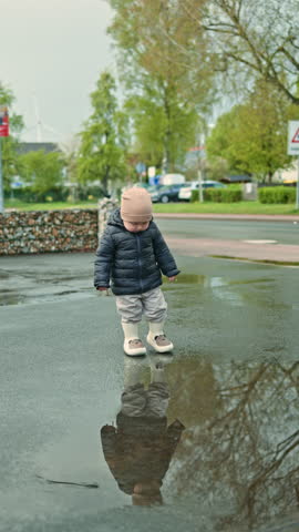 2 year old boy in rubber boots runs through puddles. Child rejoices in the rain and runs on wet asphalt. Boy on a rainy day on a walk on the street.