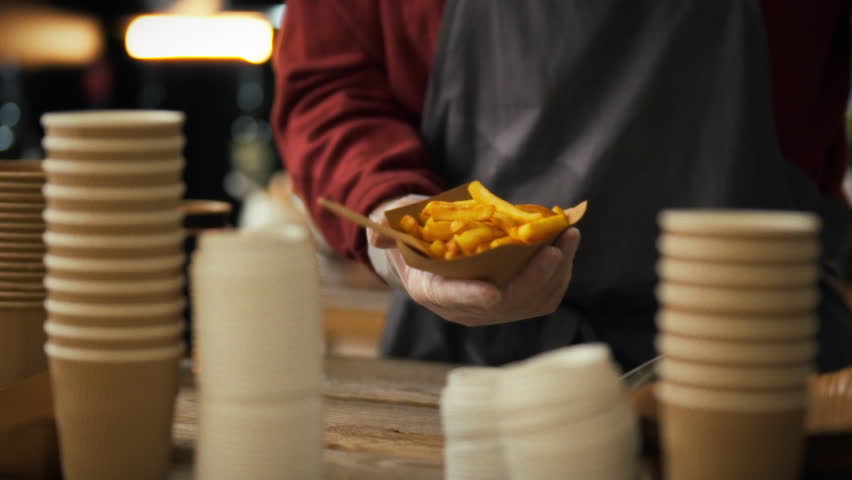 An experienced fast food cook collects orders for customers. He uses metal tongs to place fresh fried potatoes into paper packaging. Preparing food for guests of fast food establishments