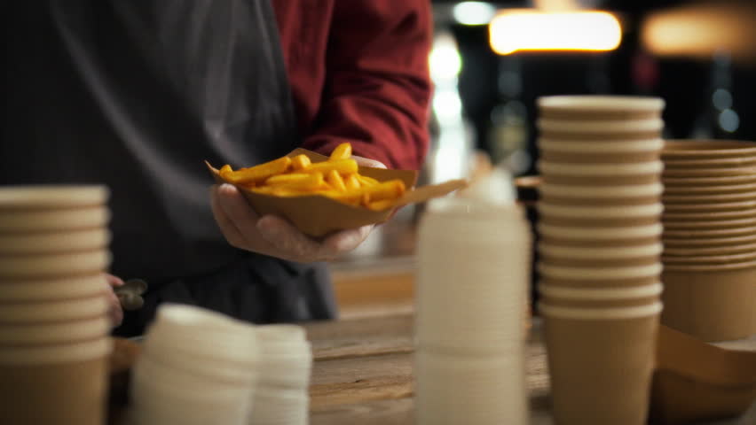 An experienced young fast food chef prepares dishes for customers. He carefully places fresh herbs on French fries with tongs. Work on assembling orders for customers of fast food establishments