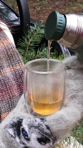 A girl's hand in a woolen mitten holds a glass cup with a double bottom against the background of a pine branch, hot tea is poured into it from a thermos