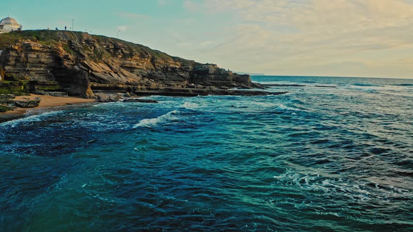 Drone flies low over the surface of the water along the rocky shore during sunset. Drone is flying along beautiful  ocean coast.   Beautiful natural landscape with ocean rocky shore.  Cinematic color