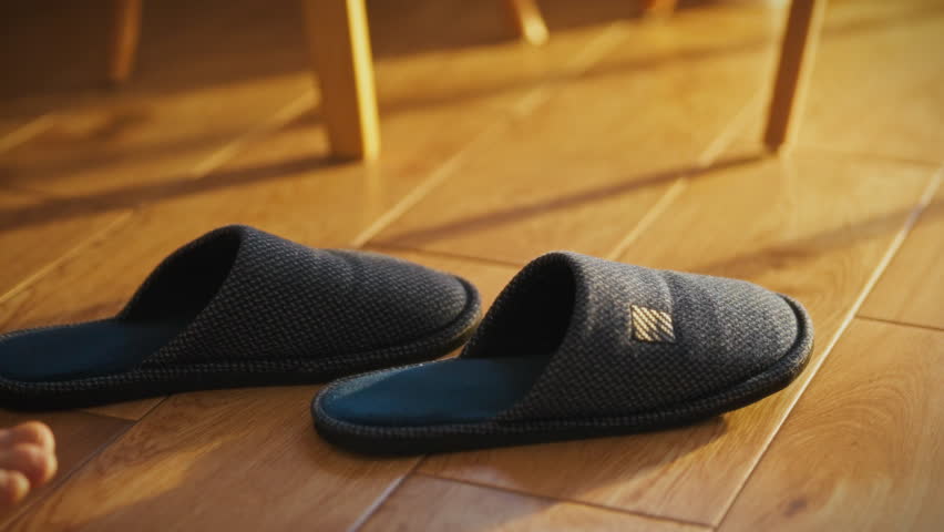 A man reaches for a pair of slippers with his foot while enjoying the morning sunlight streaming in. The setting indicates a relaxed morning routine in a comfortable home