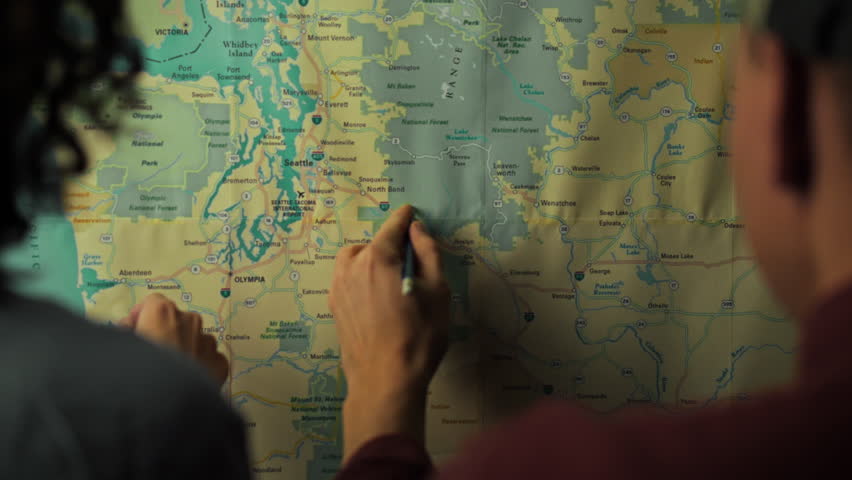 Two individuals focus on a detailed map of Washington state, marking locations and routes. This planning session takes place indoors, aiming to coordinate their upcoming journey
