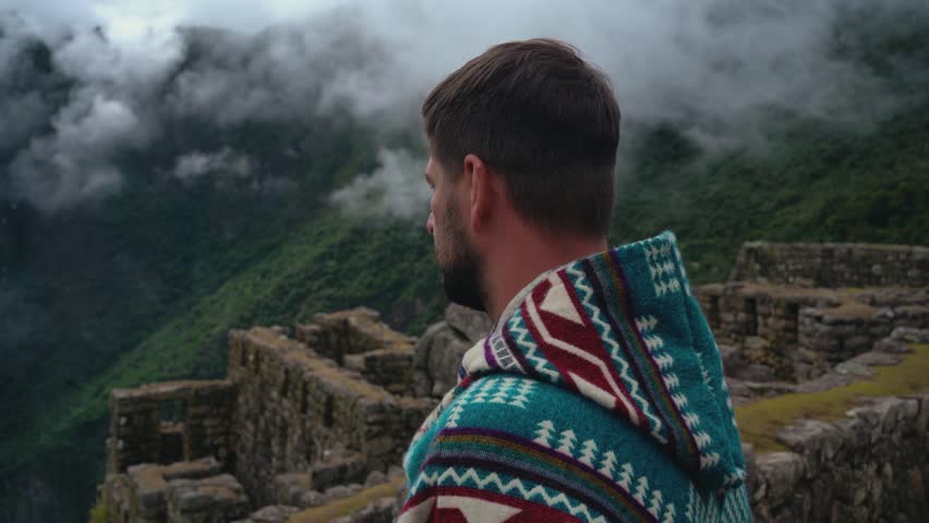 Man in Poncho Standing Amidst Machu Picchu Ruins, Gazing at Foggy Mountains