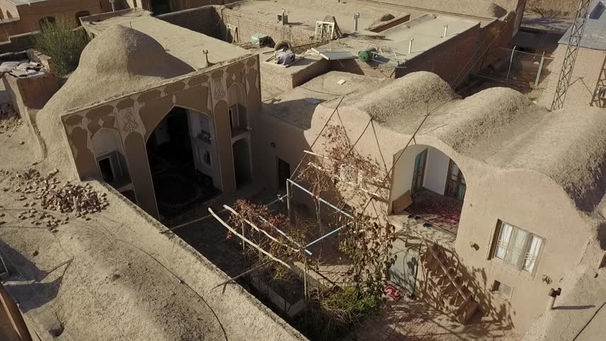Drone aerial perspective of old Persian village houses with mud rooftops, inner courtyards, and historical architecture surrounded by clay walls and narrow alleys under clear sunlight.

