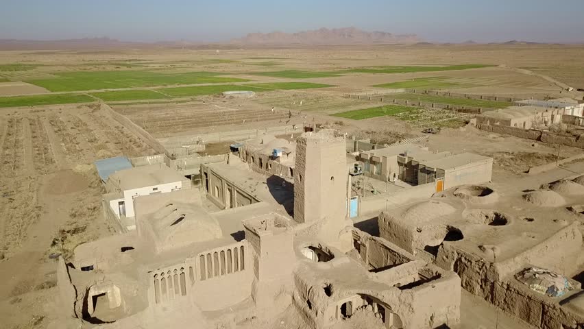 Aerial perspective of a ruined fortress and windcatcher structures surrounded by desert farms and scattered village homes. The scene depicts a blend of Persian environmental architecture, ancient 