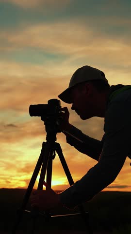 Photographer man with backpack uses camera with tripod against amazing beautiful sunset. Traveler with tripod, camera takes beautiful shots of landscape against amazing beautiful sunny sky with clouds