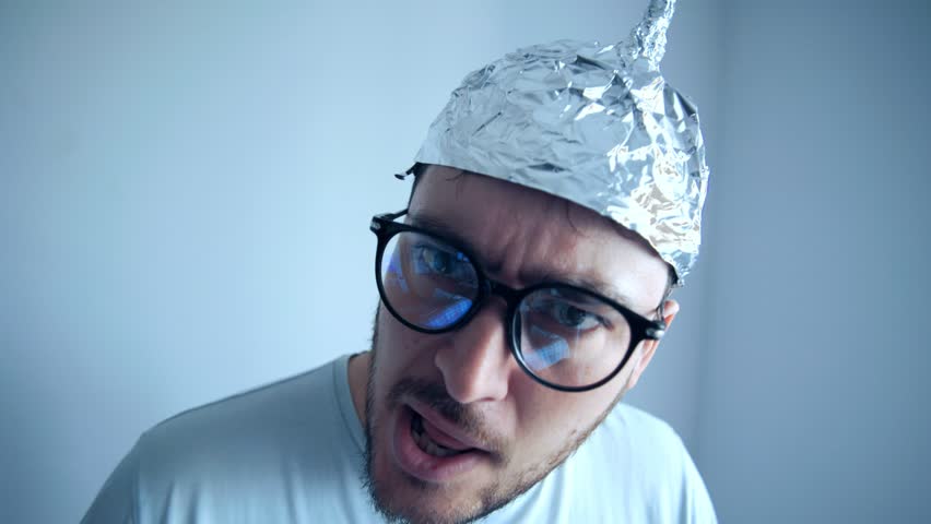 A man wearing an aluminum tinfoil hat looks skeptically at the camera with a bewildered expression. His eyeglasses amplify his humorous yet serious demeanor.