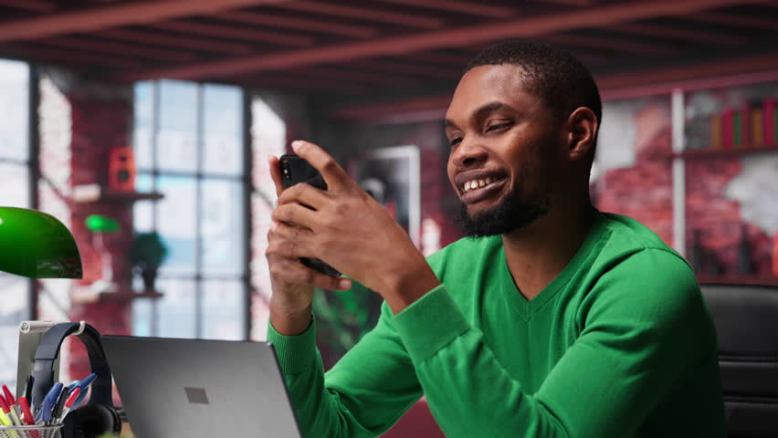 Man at home using smartphone to compose social media posts, producing content as a freelancer. African american guy monitoring profile engagement using mobile phone. Camera A.