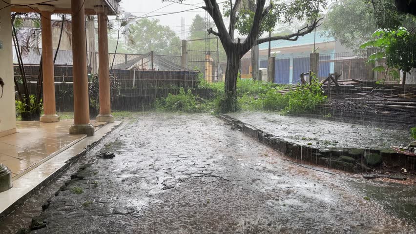 Heavy Rain Pouring Down in a Rural Yard with Lush Greenery and Weathered Structures in the Background During Daytime