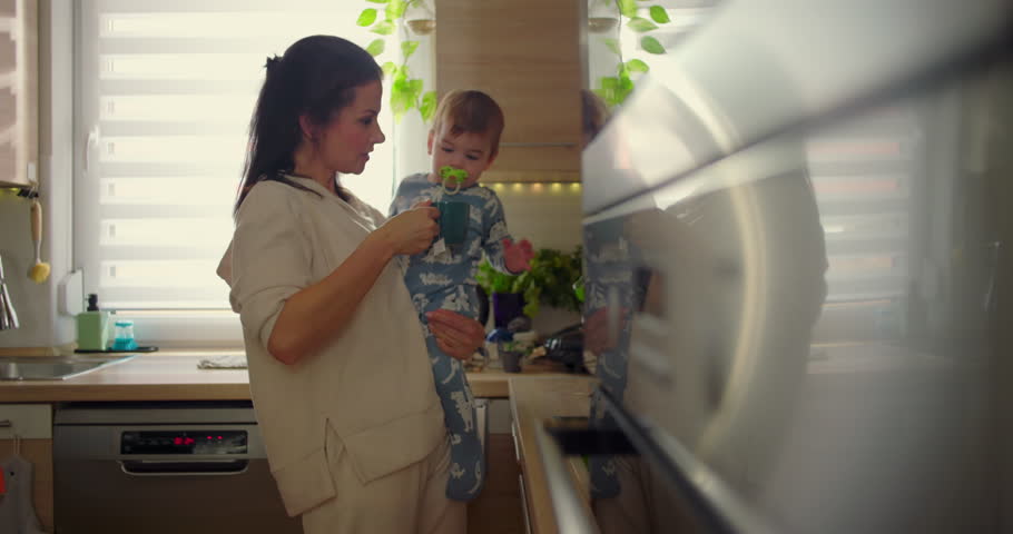 Mother holding baby in kitchen as child reaches for hot cup, protective parenting moment during everyday family routine, nurturing and safety in a warm home setting