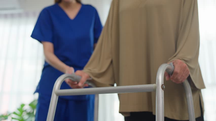 Close up of senior woman patient doing physical therapy with caregiver. Attractive specialist carer help and support elderly mature grandma practice walking slowly with walker at nursing home care.