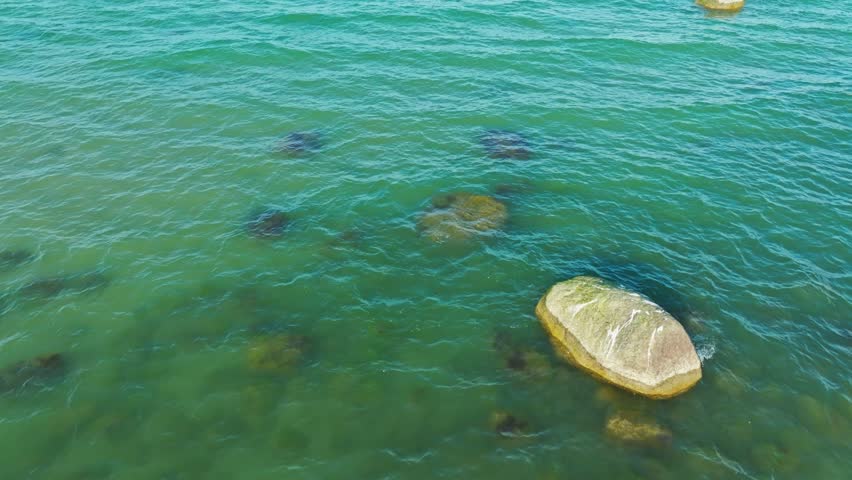 Turquoise waters gently lap against visible rocks and patches of marine vegetation, creating a serene coastal atmosphere on a clear day.