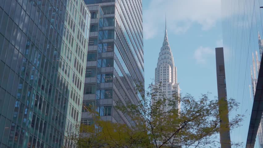 The Chrysler Building stands tall in Midtown Manhattan, seen between sleek glass buildings and city foliage under a clear sky.