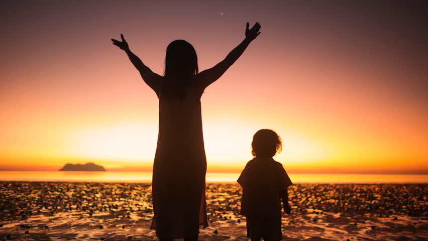 Silhouette of mother raising arms toward the sunset as child sits nearby, expressing freedom, gratitude, and presence in a serene golden hour moment by the sea.