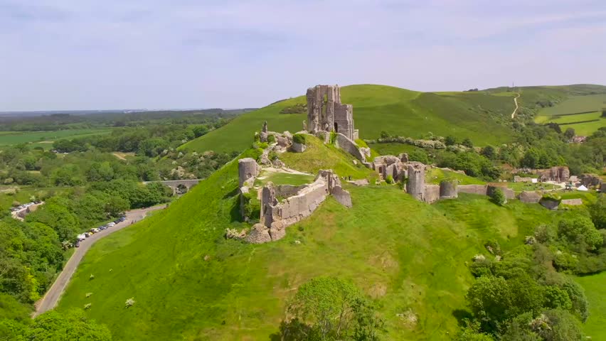 Drone pulls away from Corfe Castle ruins, revealing its hilltop location surrounded by green countryside and scenic landscapes in Dorset, southern England