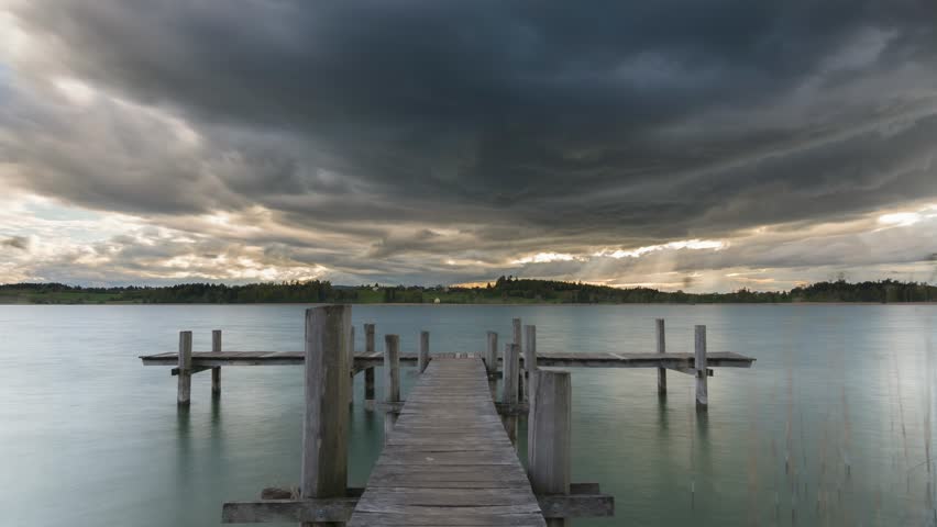 Stormy clouds move over a lake. Sunbeams break through the clouds and shine on the landscape. The storm passes. Beautiful nature, captured from a jetty. Time-lapse.