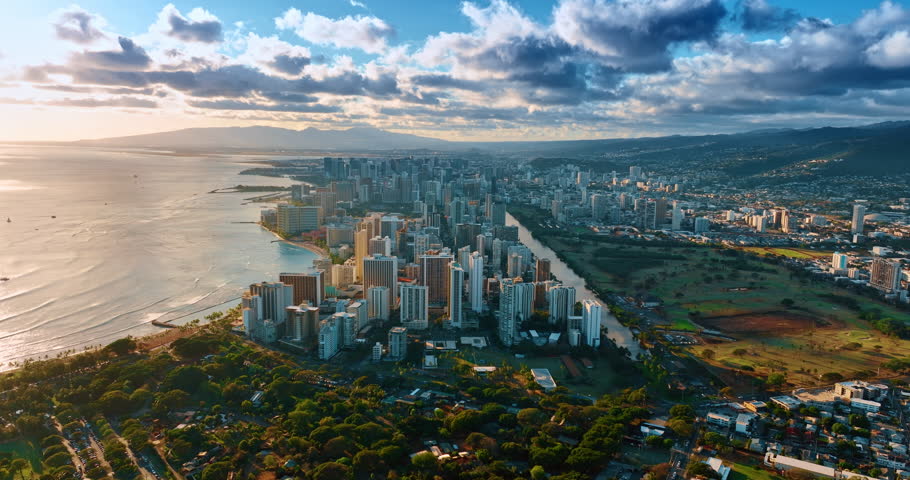 Flying closer to Waikiki resort on the shore of the Pacific Ocean in the light of setting sun. Vast urban landscape of Honolulu under the cloudy sky.