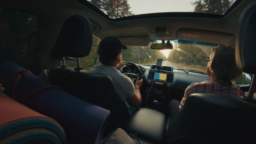 Adult man and elderly woman travel in the car on a rural road during sunset with hiking gear laying inside the car