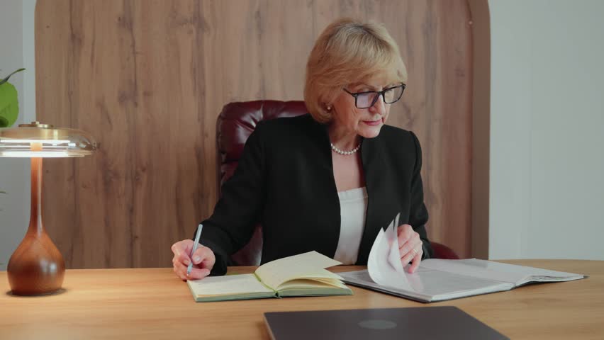 A 60-year-old woman reviews a document folder and writes notes in a notebook while the camera pans slowly from left to right. A visual of focused lifelong learning at home. 4k footage
