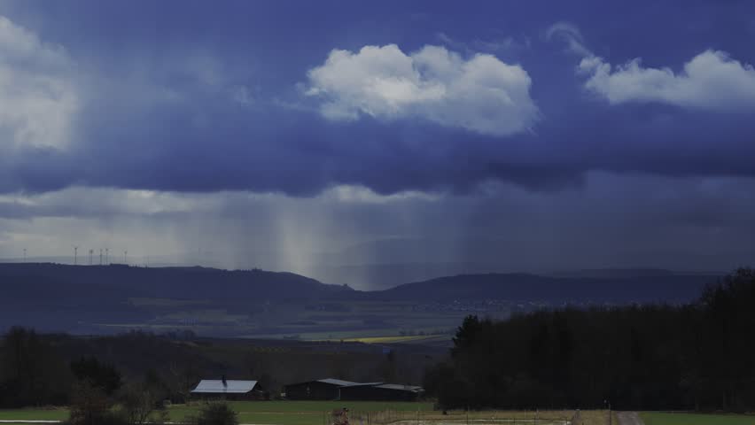 A time-lapse footage of the dense clouds moving over a rural farm with green fields at sunset, with wind farm on rolling hills on the horizon