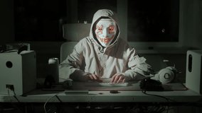 Person in LED mask and hooded jacket sitting at desk in dark gaming room, typing on keyboard. Mysterious atmosphere suggests cyberpunk hacker, gaming roleplay or digital performance art. White hacker - Powered by Shutterstock - Get 15% off with code: PIKWIZARD15