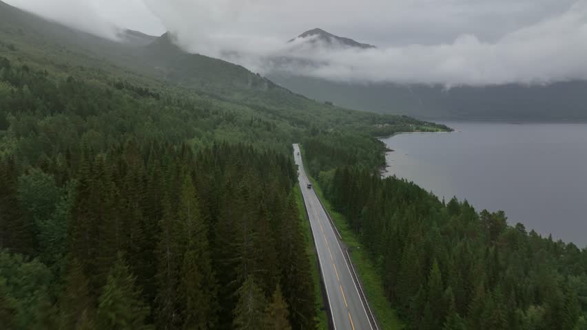 A drone view of vehicle driving on a road through hillside forest by the sea on a cloudy day in northern Norway