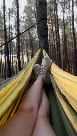 Close-up legs of female tourist swinging in hammock vertical video relaxing on vacation in pine forest in summer at sunset. Camping, hiking, healthy lifestyle, meditation, freedom. Travel concept