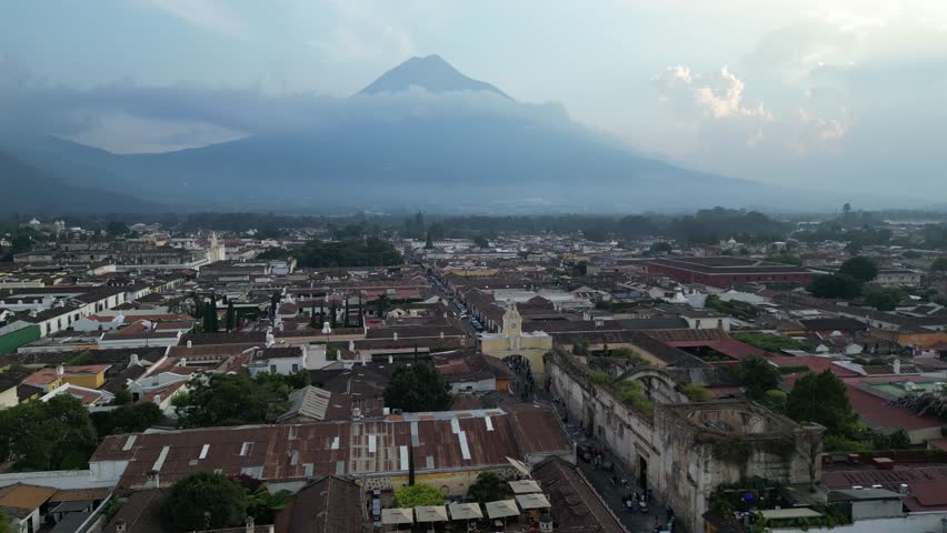 sunset aerial drone view of volcano agua in antigua guatemala with colorful colonial rooftops clouds downtown landmark arch church ruins scenic footage street flying above city dusk mountain tourism
