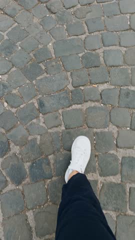 Vertical video single white sneaker on gray cobblestone pavement walk woman tourist. Person wearing dark blue pants walking. Urban exploration. City street. Shoe stepping on rough cobblestone ground