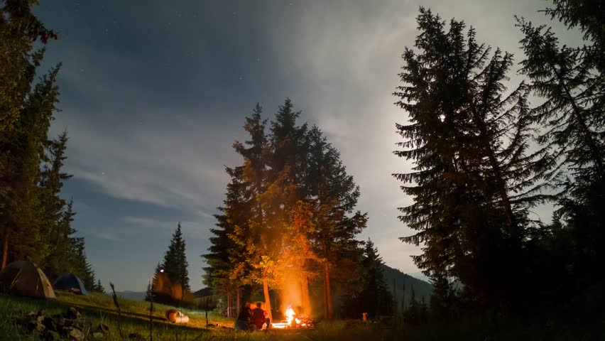 Tranquil camping scene with touristic tents nestled among tall pine trees under partly cloudy, star-studded sky. Tourists having a rest at campfire illuminates area, with light trails.