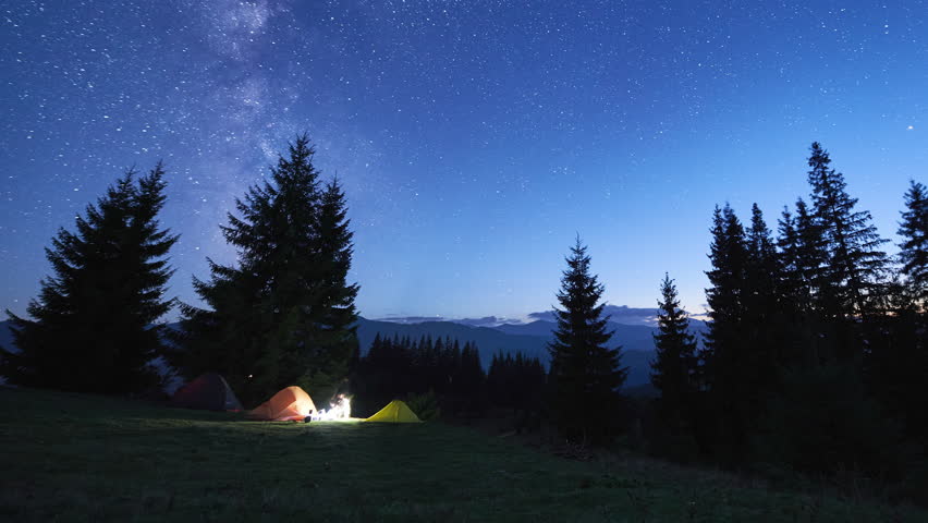 Night camping in mountains. Amidst forested mountain landscape, two tents glow warmly beside campfire under brilliant, star-filled sky. Silhouettes of tourists resting at campsite under Milky Way.