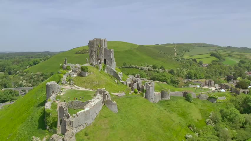 Drone image capturing Corfe Castle’s ancient ruins, rolling green hills, and nearby village activity in Dorset, England on a bright and clear summer day
