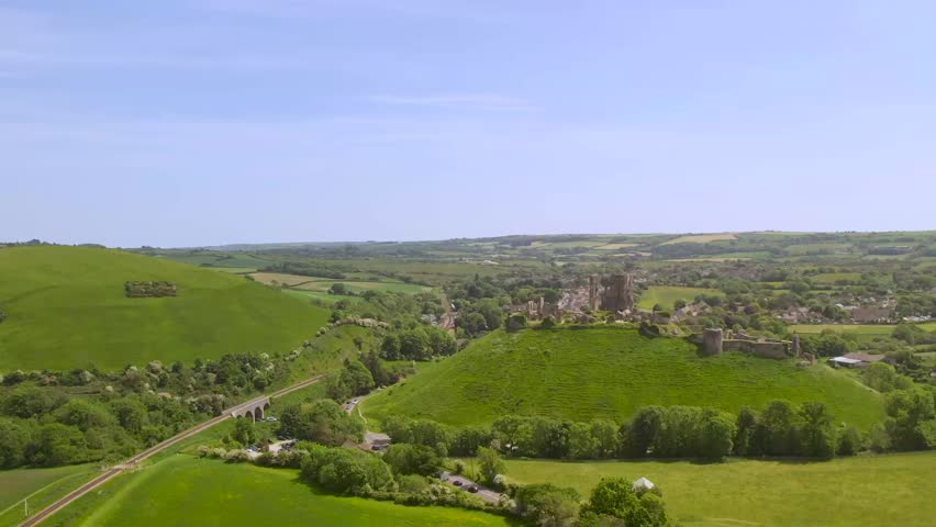 Panoramic aerial image of Corfe Castle in Dorset, England, showing the hilltop ruins, surrounding countryside, village, and railway viaduct on a sunny summer day