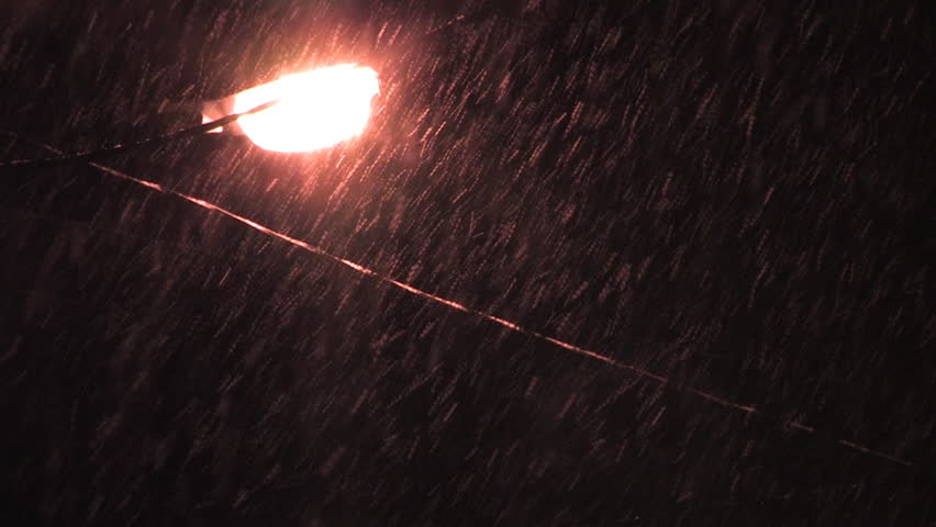 Snow falls at night near a street light.