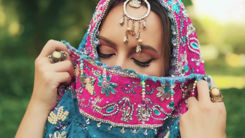 Beautiful Indian fantasy woman posing hides beauty face behind scarf, sexy eyes looking at camera festive holiday make-up. Oriental girl India fashion style. Summer garden green tree nature park