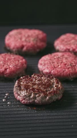 cook turn around minced beef burger meat patties cooking on hot black electric grill surface, food closeup