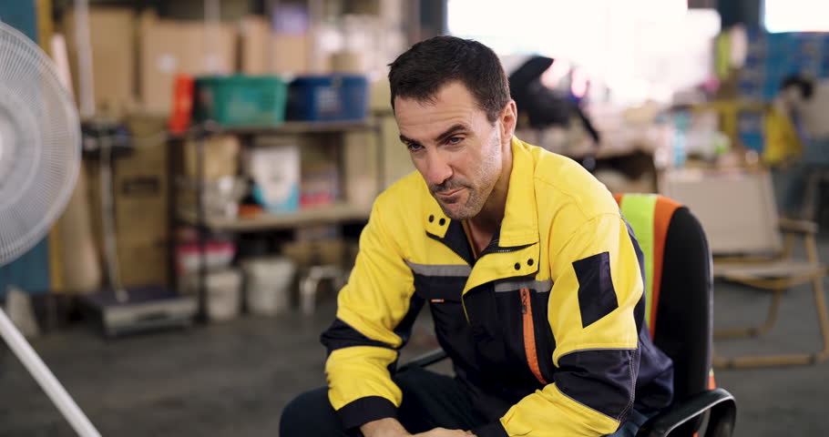 Warehouse male worker in uniform wiping sweat while sitting on chair exhausted after work task showing fatigue, stress and human side of industrial labor environment