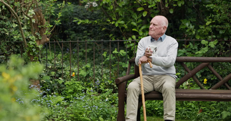 Happy senior man, coffee and park bench in garden for morning, relax or break at retirement home. Calm elderly male person drinking tea in mug or cup sitting with kane on outdoor chair in nature