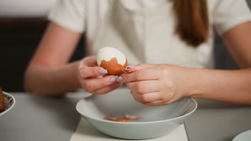 Close up of fair skinned woman peeling boiled egg over dish with egg shell pieces inside, wearing white shirt and apron, blurred view of brown hair flowing down and edge of another plate