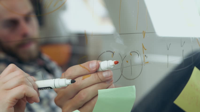 Close up of serious bearded man drawing marks on glass wall in office room to see vision of further business strategy