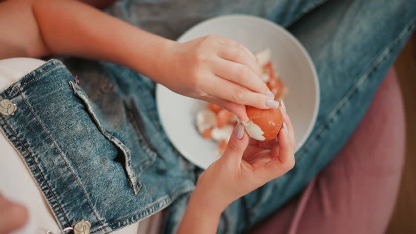 Close up of young lady in denim overall delicately removing brown egg shell into white plate resting on her lap while seated, with detailed view of polished nails and egg fragments filling the bowl