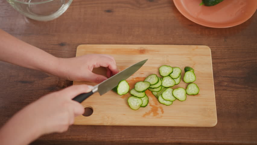 Overhead view of person using knife to slice cucumber into thin pieces on wooden cutting board with pile of cucumber slices and plate holding whole cucumber on wooden kitchen counter