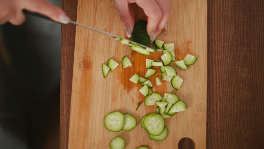 Top down view of person slicing cucumber into small pieces on wooden cutting board, with scattered cucumber slices spread across surface during meal preparation in kitchen