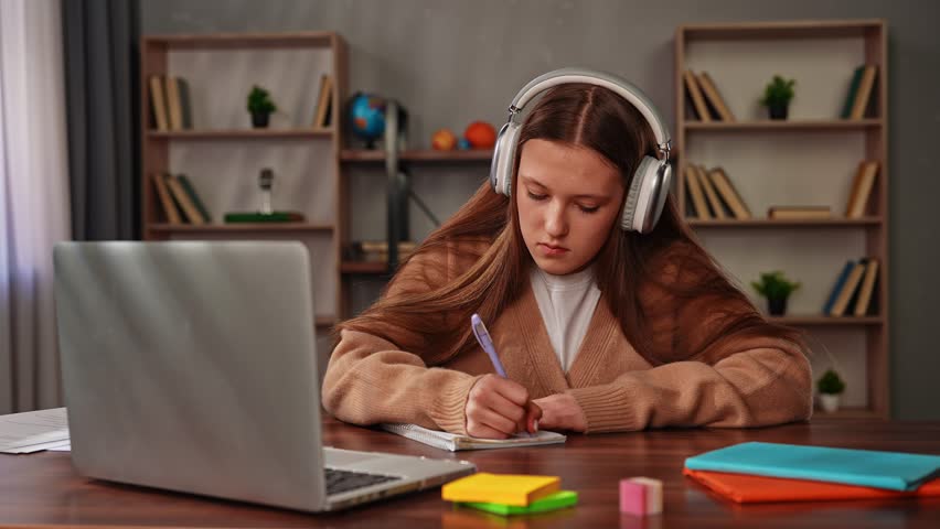 Daily lifestyle concept. Young teenage girl studying at home, girl sits at desk with laptop and books, doing homework, wearing headphones writing notes in notebook from laptop, looking at screen.