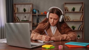 Daily lifestyle concept. Young teenage girl studying at home, girl sits at desk with laptop and books, doing homework, wearing headphones writing notes in notebook from laptop, looking at screen. - Powered by Shutterstock - Get 15% off with code: PIKWIZARD15